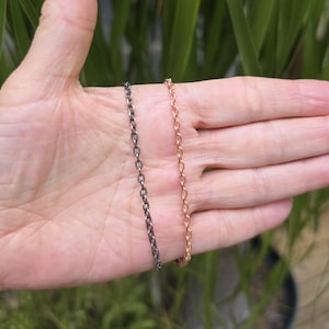 May include: Two delicate metal chains are displayed side by side. One chain features a dark, oxidized finish with a textured surface on each oval link, while the other chain has a bright, polished rose gold tone with a matching textured link design.