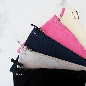 May include: Five zippered pouches in different colours, including black, grey, navy, pink, and natural. Each pouch has a zipper closure and a label with the colour name.