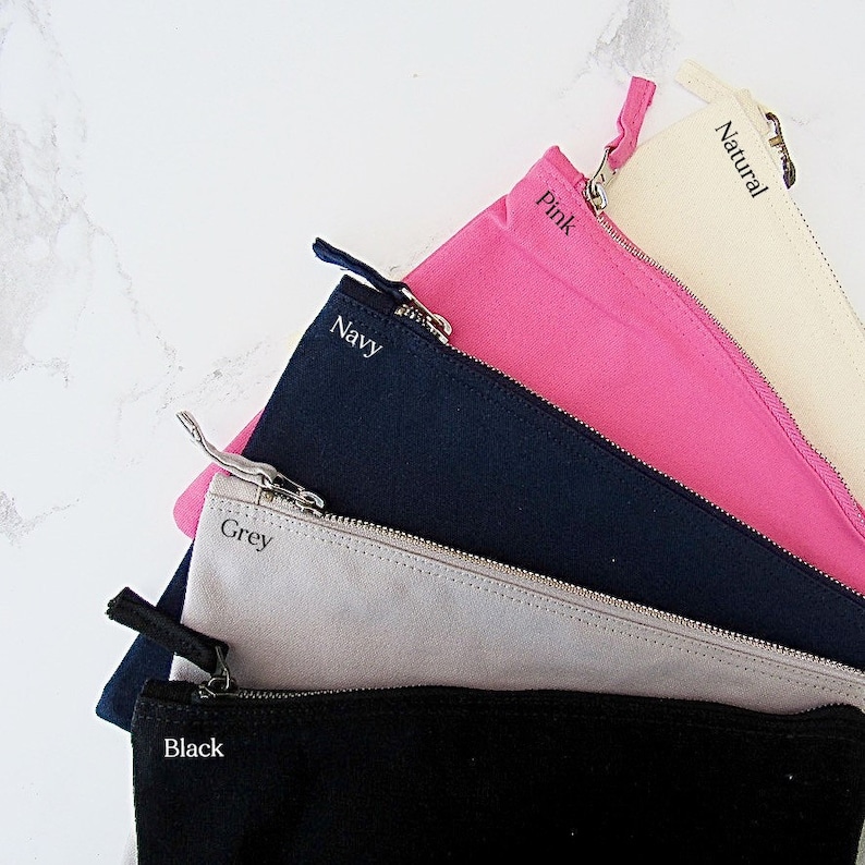 May include: Five zippered pouches in different colours, including black, grey, navy, pink, and natural. Each pouch has a zipper closure and a label with the colour name.