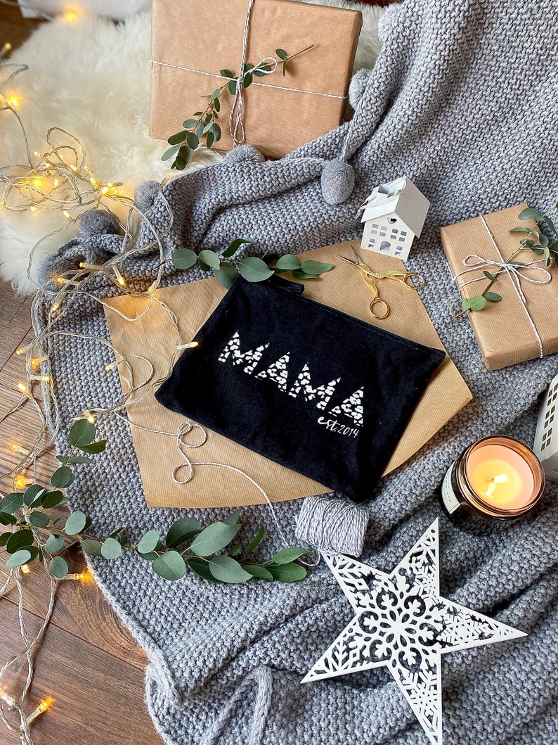 May include: A black zippered pouch with the word "MAMA" in white letters and the text "est. 2014" underneath. The pouch is surrounded by a grey knitted blanket, a white star, a lit candle, and wrapped gifts.