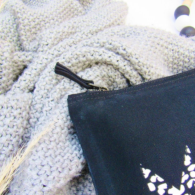 May include: A black canvas zippered pouch with a white design. The pouch is lying on a grey knitted fabric.
