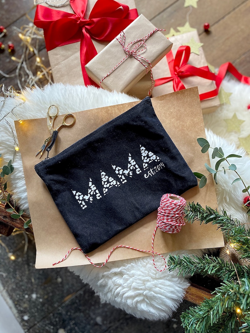 May include: Black canvas zippered pouch with the word "MAMA" in white letters with a white polka dot pattern. The text "est. 2014" is in white letters below the word "MAMA". The pouch is on a brown paper surface with red and white twine.