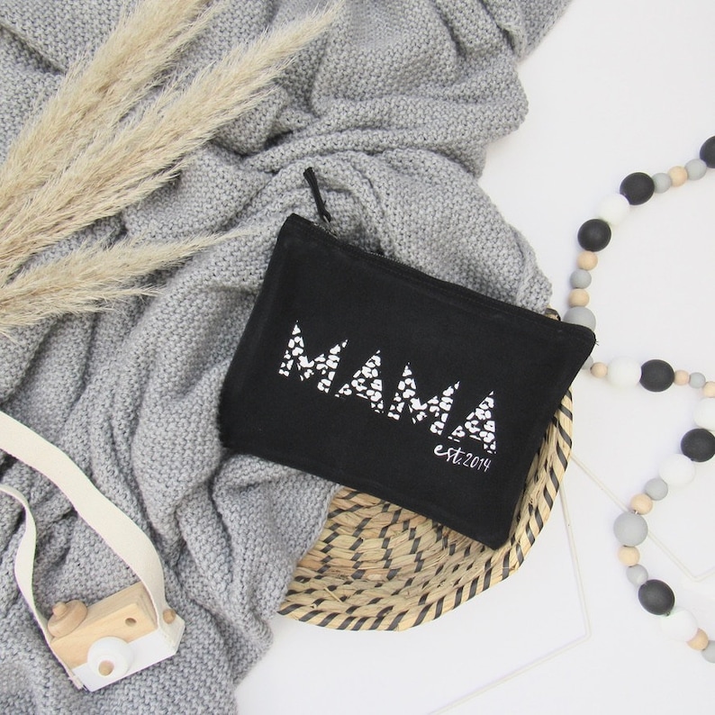 May include: Black zippered pouch with a white "MAMA est. 2014" design. The pouch is sitting on a woven basket.
