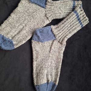 May include: A pair of hand-knitted socks. The socks are primarily gray with blue heels and toes. The cuffs are ribbed with two blue stripes. The socks are made of a soft, textured yarn.