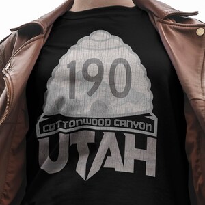 May include: A black t-shirt with a white graphic design featuring a stylized road sign with the number 190 and the text "Cottonwood Canyon Utah".
