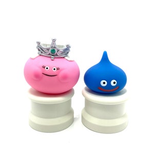 Puede incluir: Dos figuras de personajes de dibujos animados. Una es rosa con una corona plateada y la otra azul. Ambas tienen rasgos faciales simples y están sobre bases cilíndricas blancas. El fondo es blanco.