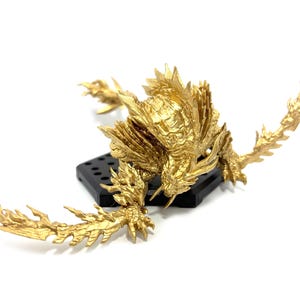 May include: A gold-colored figurine of a dragon-like creature with intricate details. The figure is posed on a black base, with outstretched wings and a textured, scaled appearance. The creature is a vibrant gold color.
