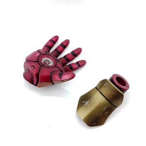 May include: A red and gold plastic action figure hand and forearm. The hand is open and the forearm is closed. The hand has a red and black design and a clear plastic circle in the center. The forearm is gold with a red plastic ring at the top.