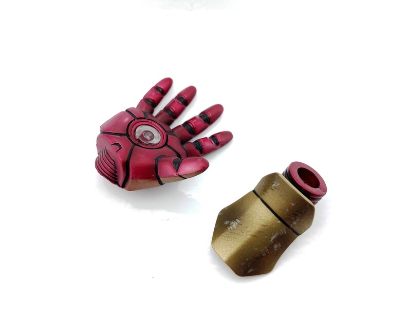 May include: A red and gold plastic action figure hand and forearm. The hand is open and the forearm is closed. The hand has a red and black design and a clear plastic circle in the center. The forearm is gold with a red plastic ring at the top.