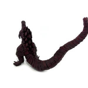 May include: A maroon-colored Godzilla figurine, viewed from the back, with a long, textured tail. The figure is set against a white background, highlighting the details of the creature's dorsal fins and scaly skin.