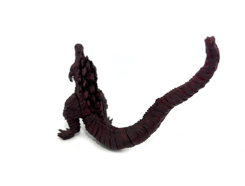 May include: A maroon-colored Godzilla figurine, viewed from the back, with a long, textured tail. The figure is set against a white background, highlighting the details of the creature's dorsal fins and scaly skin.