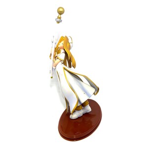 May include: A figurine of a character in a white and gold outfit, holding a staff. The figure has long blonde hair and is standing on a brown oval base. A golden orb is detached above the figure.