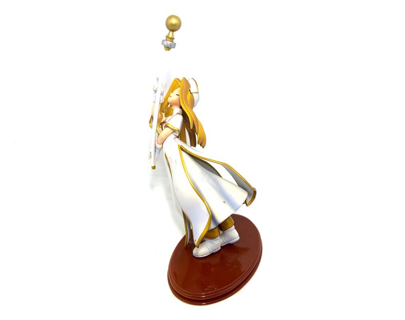 May include: A figurine of a character in a white and gold outfit, holding a staff. The figure has long blonde hair and is standing on a brown oval base. A golden orb is detached above the figure.