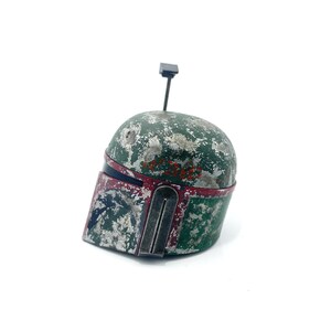 May include: A weathered green and red Boba Fett helmet with a black antenna on top. The helmet has a worn and distressed look, with visible scratches and paint chips.