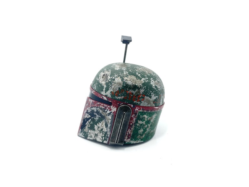May include: A weathered green and red Boba Fett helmet with a black antenna on top. The helmet has a worn and distressed look, with visible scratches and paint chips.
