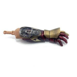 K&ouml;nnte beinhalten: Ein detaillierter Actionfigurenarm, mit einem hellbraunen Oberarm und einem metallischen Unterarm und Hand. Der Unterarm ist eine Mischung aus Silber, Gold und Rot, mit aufwendigen mechanischen Details. Die Hand ist tiefrot.