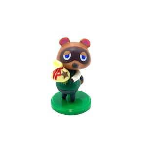May include: A green and brown figurine of Tom Nook from the Animal Crossing video game series. He is holding a yellow bag with a red star on it. The figurine is standing on a green base.
