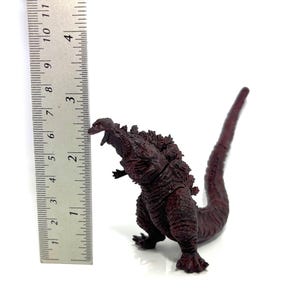 May include: A dark red Godzilla figurine, approximately 6 inches tall, with detailed textured skin and dorsal fins. The figure is posed with its mouth open and tail extended, set against a white background.