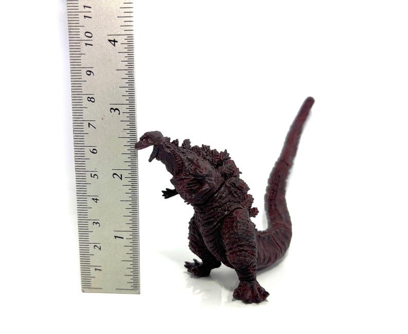 May include: A dark red Godzilla figurine, approximately 6 inches tall, with detailed textured skin and dorsal fins. The figure is posed with its mouth open and tail extended, set against a white background.