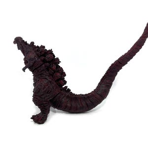 May include: A maroon Godzilla figurine with a textured, scaly appearance. The figure is posed with its mouth open and tail extended. The figurine is a collectible item.