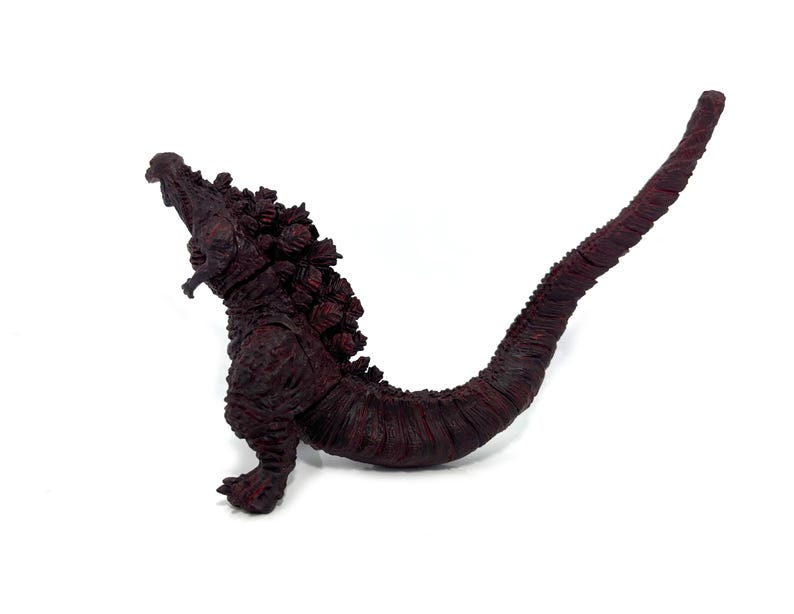 May include: A maroon Godzilla figurine with a textured, scaly appearance. The figure is posed with its mouth open and tail extended. The figurine is a collectible item.