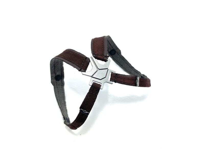 May include: A brown and gray harness with a silver metal center piece. The harness has adjustable straps and is designed to fit around the chest and shoulders. The harness is made of a durable material.