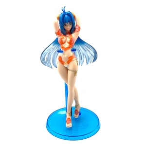 May include: A blue-haired figurine of a woman wearing an orange and gold bikini, standing on a blue base. The figurine is posed with her arms raised above her head.