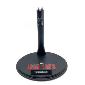 K&ouml;nnte beinhalten: Schwarzer Plastik-Actionfigurenst&auml;nder mit runder Basis. Die Basis tr&auml;gt das Marvel-Logo und den Text "IRON MAN 3" mit dem Text "THE MANDARIN" darunter.
