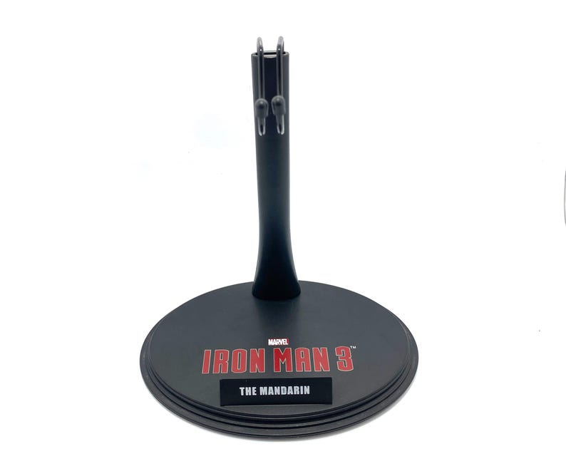 K&ouml;nnte beinhalten: Schwarzer Plastik-Actionfigurenst&auml;nder mit runder Basis. Die Basis tr&auml;gt das Marvel-Logo und den Text "IRON MAN 3" mit dem Text "THE MANDARIN" darunter.
