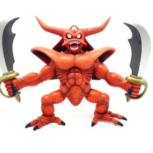 May include: A red, horned demon action figure with a third eye, holding two silver and gold curved swords. The figure has a muscular physique and is posed with its legs spread and arms outstretched.
