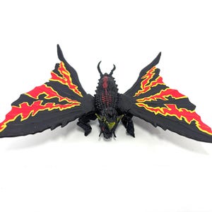May include: A black creature figurine with large, moth-like wings featuring jagged red and yellow patterns. The body is textured with dark scales, small horns on the head, and bright yellow eyes. The figure is posed with wings spread wide.