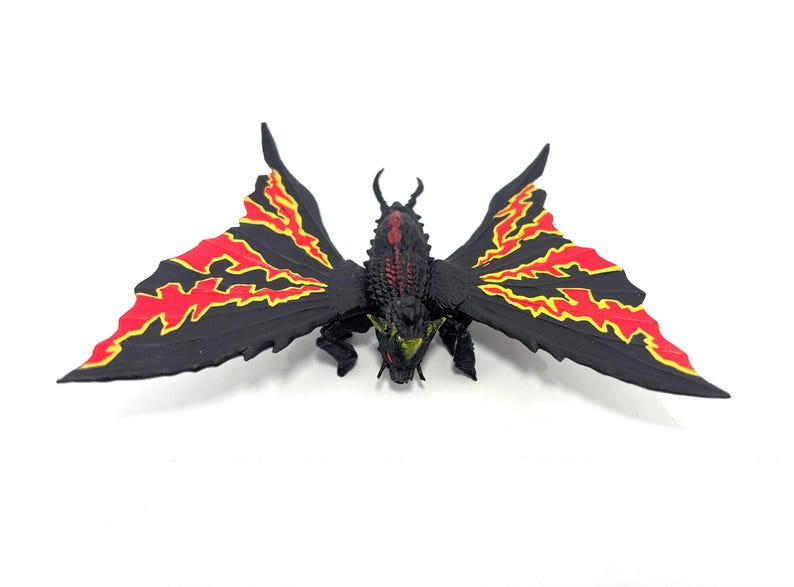 May include: A black creature figurine with large, moth-like wings featuring jagged red and yellow patterns. The body is textured with dark scales, small horns on the head, and bright yellow eyes. The figure is posed with wings spread wide.