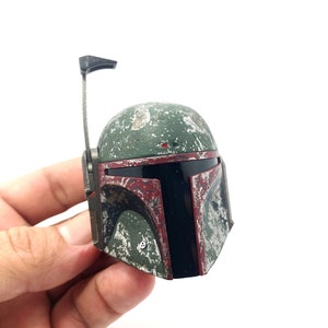 May include: A green and red plastic Boba Fett helmet with a weathered look. The helmet has a black visor and a small antenna on top.