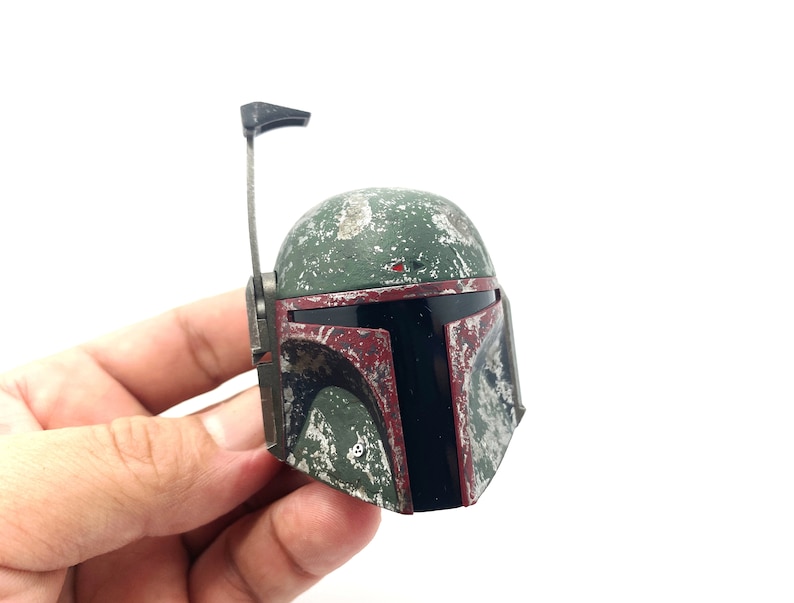 May include: A green and red plastic Boba Fett helmet with a weathered look. The helmet has a black visor and a small antenna on top.