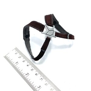 May include: A brown and gray harness with a silver metal centerpiece. The harness has adjustable straps and is designed for a small pet. A ruler is placed next to the harness for scale.
