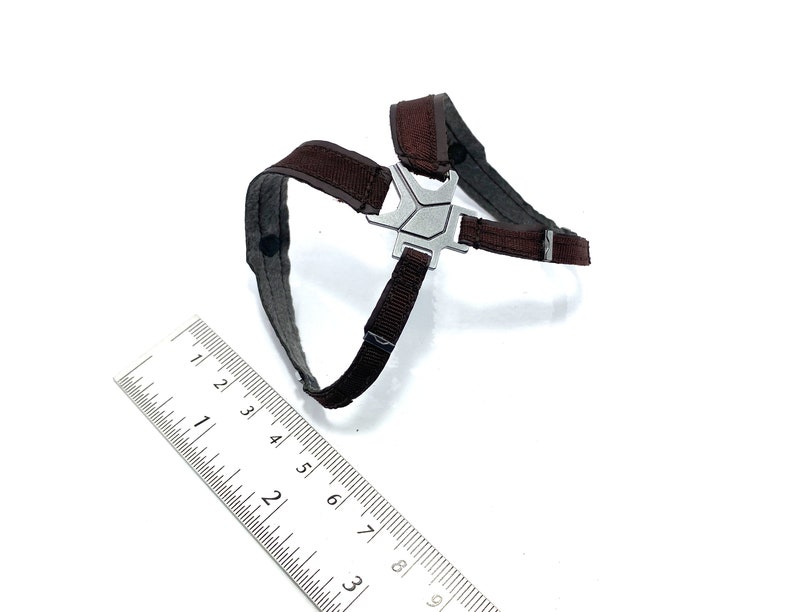 May include: A brown and gray harness with a silver metal centerpiece. The harness has adjustable straps and is designed for a small pet. A ruler is placed next to the harness for scale.