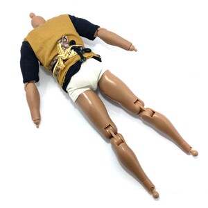 May include: A brown-skinned action figure with black and brown clothing and a white undergarment. The figure has articulated limbs and is lying on its back.