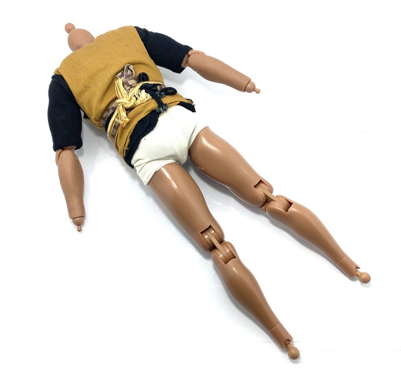 May include: A brown-skinned action figure with black and brown clothing and a white undergarment. The figure has articulated limbs and is lying on its back.