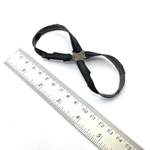 以下が含まれることがあります： 黒い生地のアクセサリー、シルバーカラーの金属アクセント付き。ベルトまたはハーネスの可能性があります。スケールとして金属製の定規が添えられています。アクセサリーの長さは約15cmです。