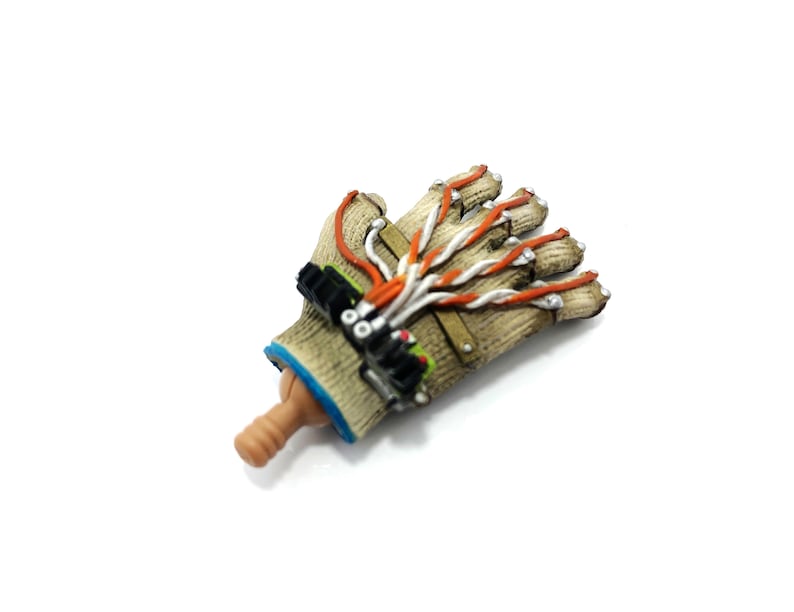 Puede incluir: Un guante marr&oacute;n y beige con cables naranja y blanco unidos a &eacute;l. El guante tiene una superficie texturizada y un pu&ntilde;o azul. El guante est&aacute; unido a una mano marr&oacute;n y beige.