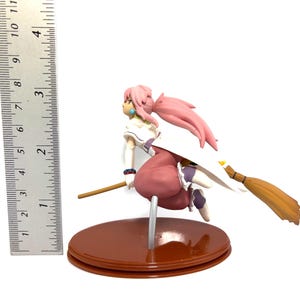 Op de afbeelding: Een beeldje van een personage met roze haar en een witte en roze outfit, rijdend op een bezem. Het beeldje staat op een bruine basis en is ongeveer 10 cm hoog. De bezem is bruin en geel.