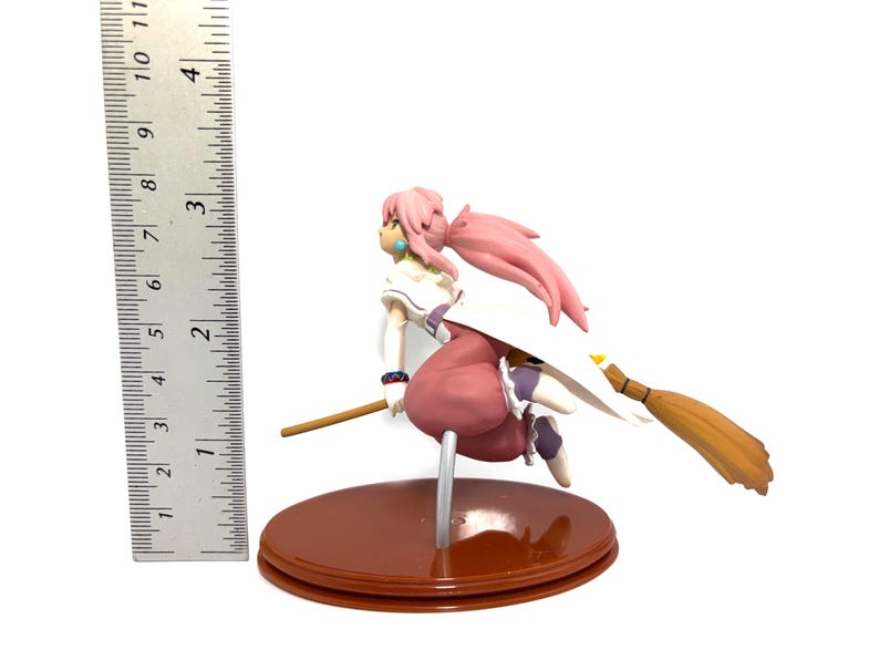 Op de afbeelding: Een beeldje van een personage met roze haar en een witte en roze outfit, rijdend op een bezem. Het beeldje staat op een bruine basis en is ongeveer 10 cm hoog. De bezem is bruin en geel.
