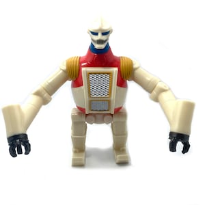 Puede incluir: Un juguete robot vintage con cuerpo color crema, pecho rojo y detalles amarillos. El robot tiene una cabeza blanca con detalles azules y manos negras en forma de garra. Se mantiene sobre dos piernas con los brazos extendidos.