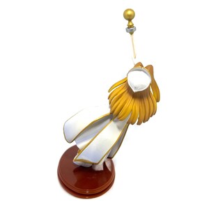 May include: A figurine of a character with golden hair and a white outfit with gold trim. The figure is mounted on a dark brown circular base and holds a golden staff with a sphere on top.