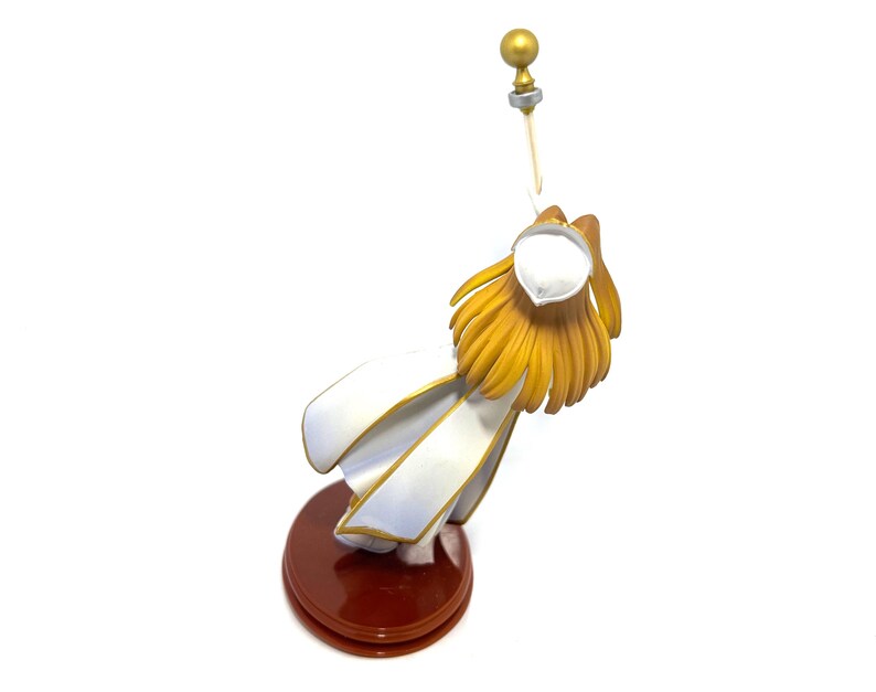May include: A figurine of a character with golden hair and a white outfit with gold trim. The figure is mounted on a dark brown circular base and holds a golden staff with a sphere on top.