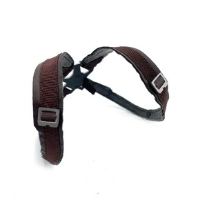 May include: A brown harness with a black plastic connector. The harness has two adjustable straps with silver-colored metal buckles. The straps are made of a woven material and are padded for comfort.