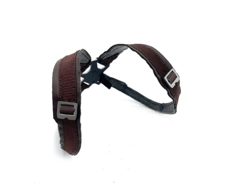 May include: A brown harness with a black plastic connector. The harness has two adjustable straps with silver-colored metal buckles. The straps are made of a woven material and are padded for comfort.