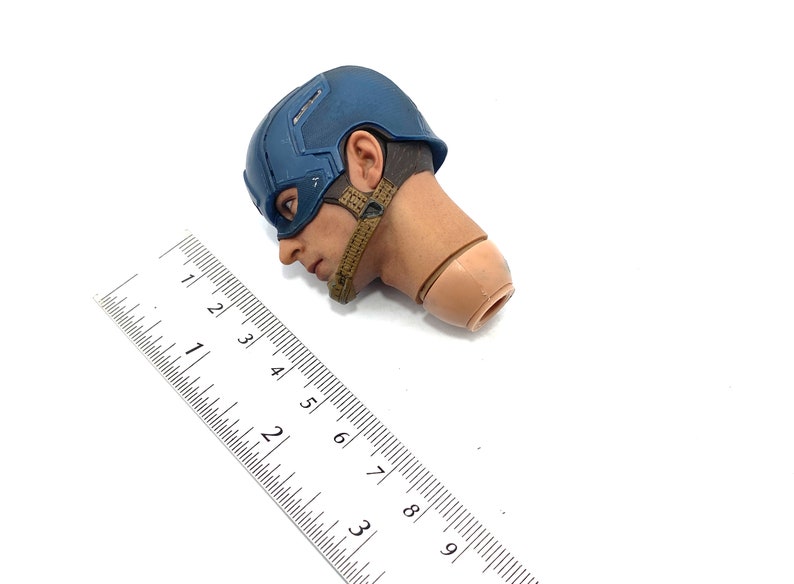 Op de afbeelding: Een Captain America actiefiguur hoofd, met een blauwe helm en een bruine riem. Naast het hoofd ligt een liniaal met centimeter metingen.