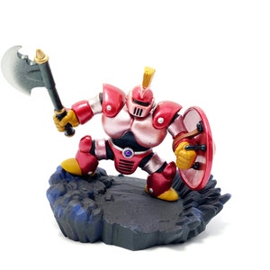 以下が含まれることがあります： 斧と盾を持った赤と銀のロボットのフィギュア。青いアクセントのある黒い岩の台座の上に立っている。