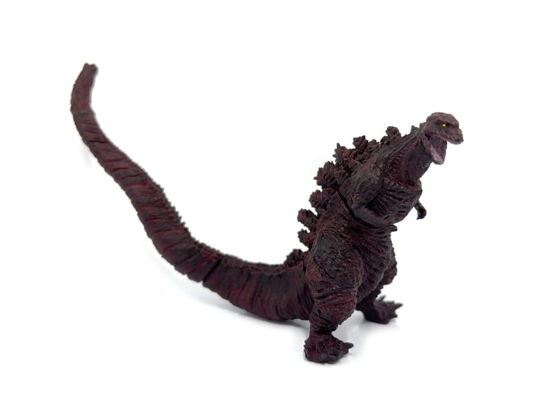 May include: A maroon Godzilla action figure with a long tail and dorsal fins. The figure is in a dynamic pose with its mouth open, showcasing detailed textures and a menacing expression. The figure is on a white background.
